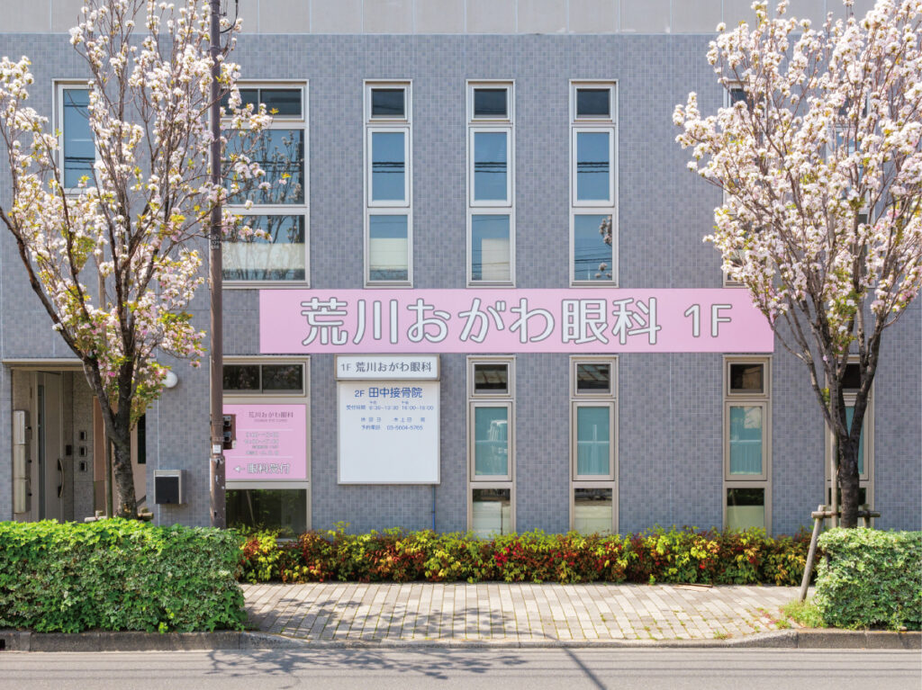荒川おがわ眼科様の外観写真。ピンク色の外看板と道沿いの桜が優しい印象を与える。